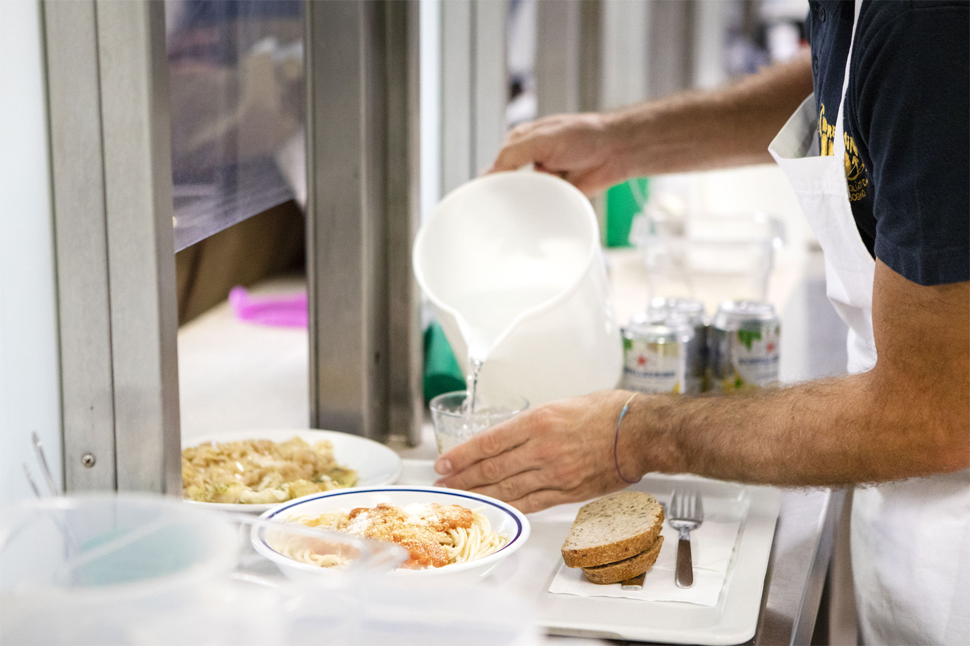 Un volontario in Cucine Popolari a Padova versa dell'acqua su un vassoio della mensa con piatti di pasta e pane